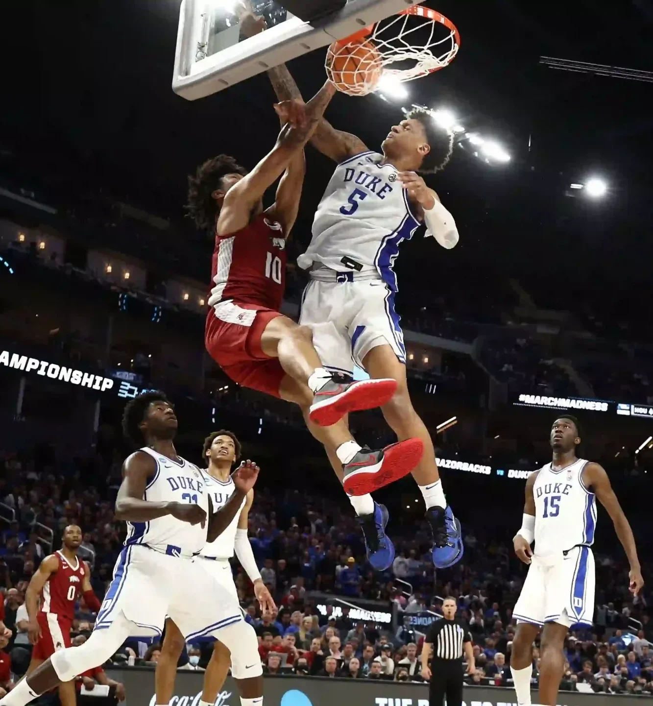 ncaa-march-madness-basketbalseizoen-amerika Een spectaculaire slam dunk van een Duke University basketballer over een verdediger van Arkansas tijdens het nationale NCAA March Madness toernooi in een vol stadion.