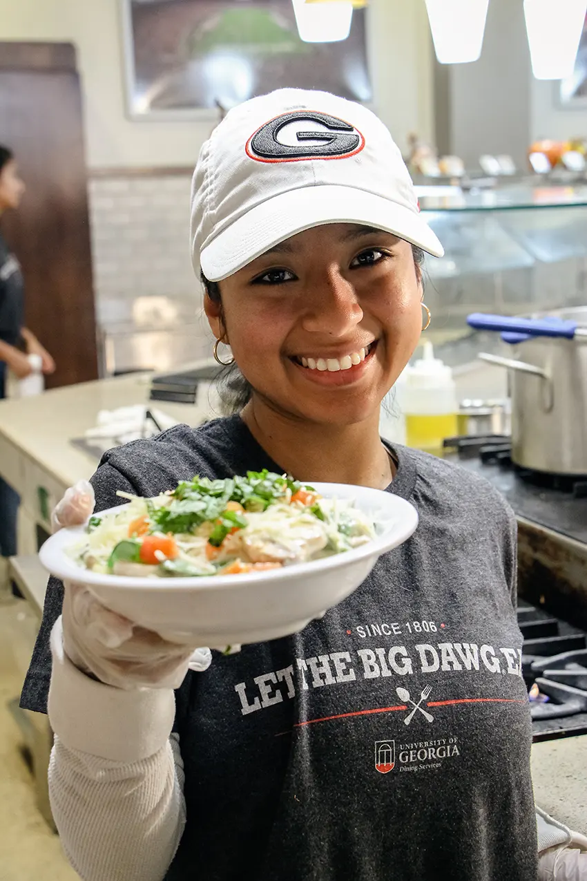 Een lachende studente van de University of Georgia, met een petje op, biedt een kom eten aan. Ze draagt een t-shirt van 'University of Georgia Dining Services