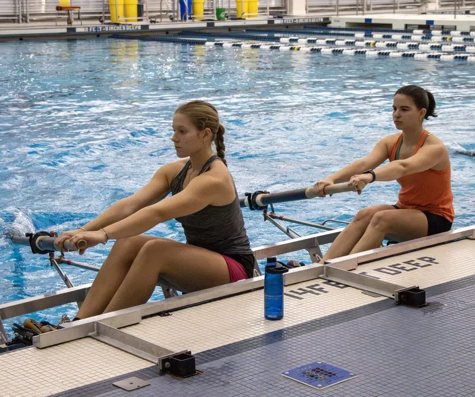 Dagelijkse training voor college roeiers Twee jonge roeisters trainen samen in een indoor roeibassin.
